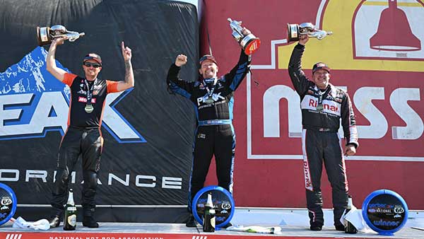Three race car drivers stand on a podium celebrating their victories. The center driver, wearing a black and blue PEAK racing suit, stands on the top step with both arms raised. The driver to the left, in a black and red Mobil 1 suit, stands on the second step holding a trophy overhead. The driver on the right, in a black and white Rinnai suit, also raises a trophy while smiling on the third step. A backdrop behind them displays the PEAK and Mission Foods logos.